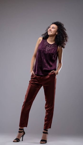 Happy relaxed young woman with curly dark hair and a lovely smile standing looking up with her hands in her pockets in trendy pants and stilettos over grey - Foto, Bild