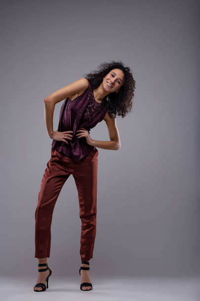 Happy vivacious young woman with a beaming smile posing bending to the side with her hands to her slim waist in stilettos and stylish pants over grey with copy space - Fotoğraf, Görsel