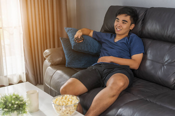 cheerful young man holding remote control and watching TV while sitting on sofa in the living room  - Photo, Image
