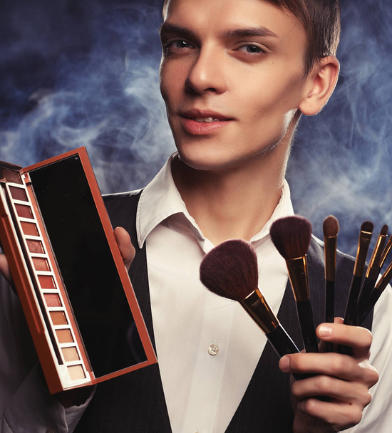 Professional makeup artist posing in studio, holds a palette of shadows and brush.Against the background of smoke. - Zdjęcie, obraz