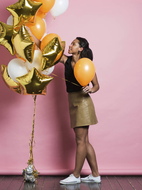 Vista lateral de la alegre joven afroamericana en falda de cuero de moda y zapatillas de deporte tomando globo naranja brillante de colorido manojo colgando cerca y sonriendo de pie sobre fondo de estudio rosa
 - Foto, imagen