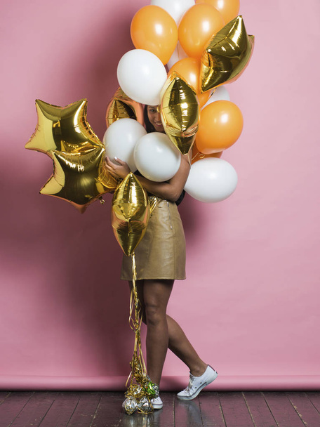 Side view of happily smiling young African American woman peeking out from large bunch of assorted bright balloons held in hands and looking at camera standing on pink background - 写真・画像