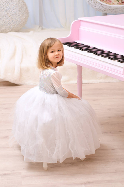 nice girl in white gown with tutu sitting at opened pink grand piano indoors - Photo, Image