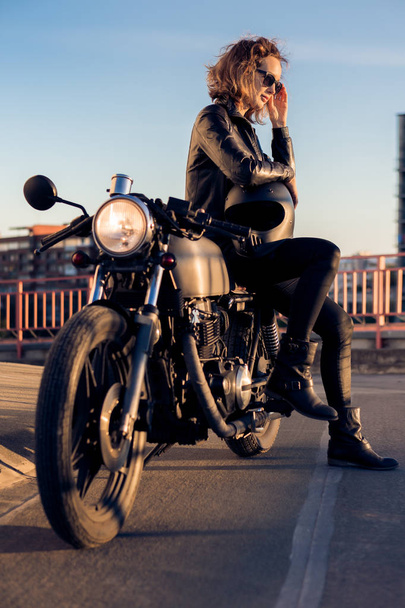 Sexy biker woman in black leather jacket sit on vintage custom caferacer motorcycle and correct sunglasses. Urban roof parking, sunset in big city. Traveling and active hipster lifestyle. Girls power. - Fotoğraf, Görsel