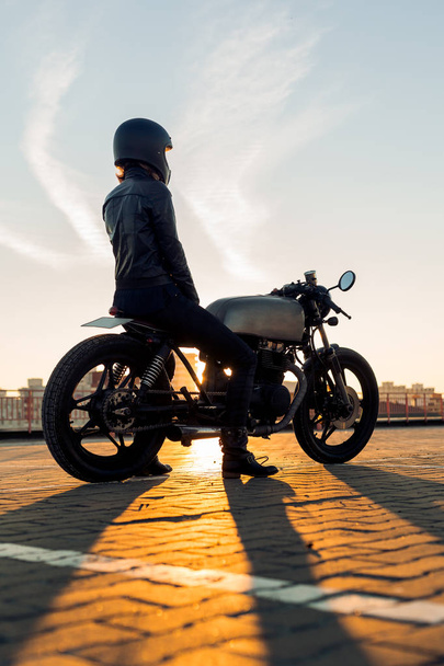 Sexy biker female in black leather jacket and full face helmet sit on vintage custom caferacer motorbike. Urban roof parking, sunset in big city. Traveling and active hipster lifestyle. Girls power. - Photo, image