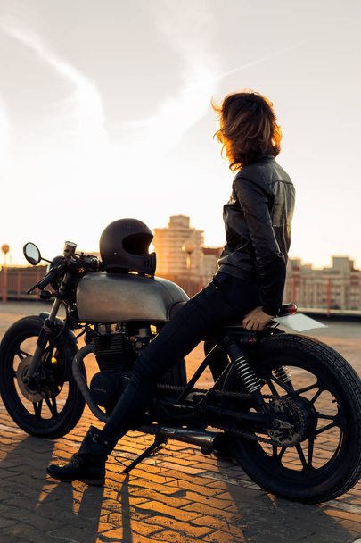 Sexy biker woman in black leather jacket sit on vintage custom caferacer motorbike and touch sunglasses. Urban roof parking, sunset in big city. Traveling and active hipster lifestyle. Girls power. - Foto, afbeelding