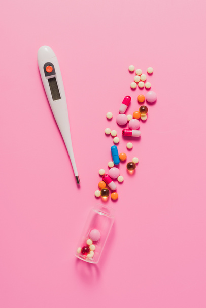 top view of various spilled pills from bottle and electric thermometer on pink - Foto, imagen