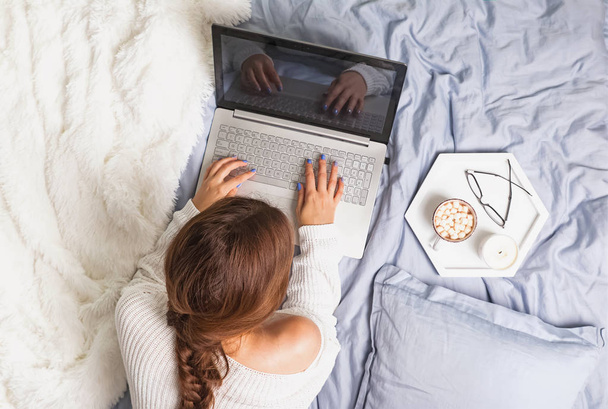 Young woman in white sweater lying on the bed and using laptop - Foto, Imagem
