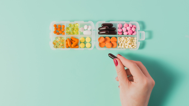 cropped shot of woman taking capsule from plastic case with various sorted pills on blue - Фото, изображение