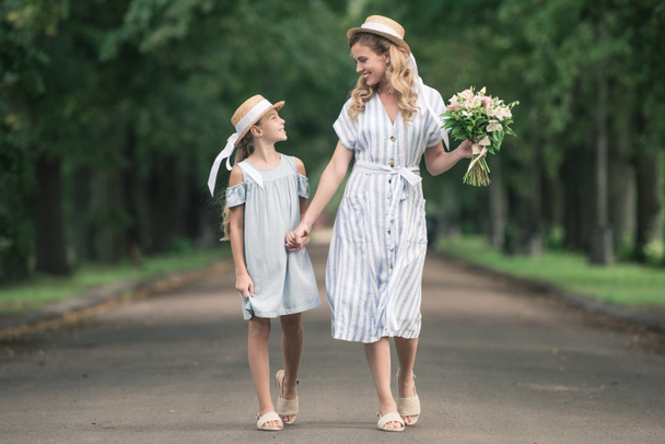 mother and daughter in straw hats holding hands and walking in green park - Photo, Image