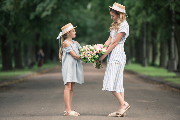 mom and daughter in straw hats with flower bouquets holding hands in park - Photo, Image