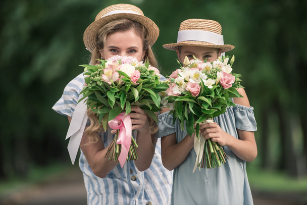 happy mother and daughter in straw hats posing with flower bouquets in front of the faces - Фото, изображение