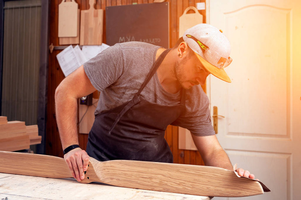 Рабочий в кепке и рубашке полирует деревянный блок наждачной бумагой перед покраской в мастерской, на заднем плане, инструментами и сверлильной машиной
.  - Фото, изображение