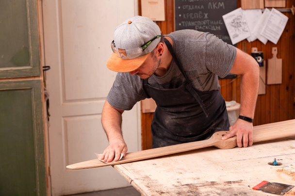 Ein Arbeiter in Mütze und Hemd poliert den Holzblock mit Schleifpapier, bevor er in der Werkstatt malt, im Hintergrund Werkzeuge und Bohrmaschine.  - Foto, Bild