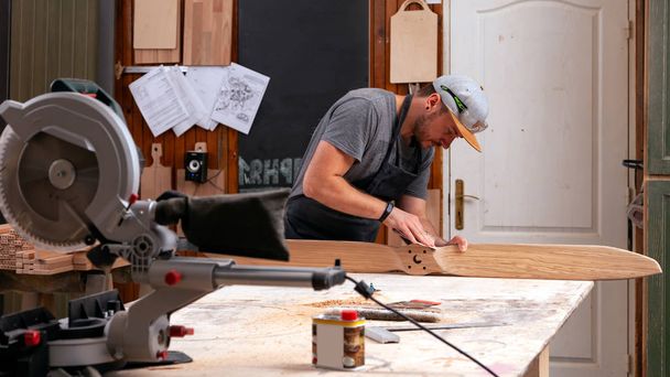 Tapasztalt asztalattal rendelkező asztalatos munkaruha és kisvállalkozás tulajdonos dolgozik fafeldolgozó műhelyben, csiszolópapírt használ gyermekhelikopter pengék csiszolásához munkaasztalnál műhelyben   - Fotó, kép