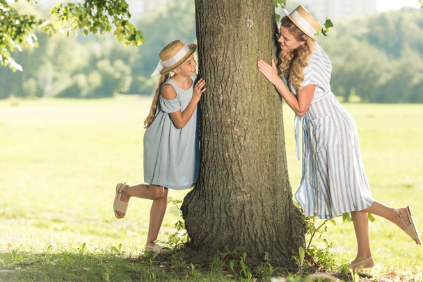 attractive mother and happy daughter in straw hats posing near tree on green lawn - Foto, Imagen