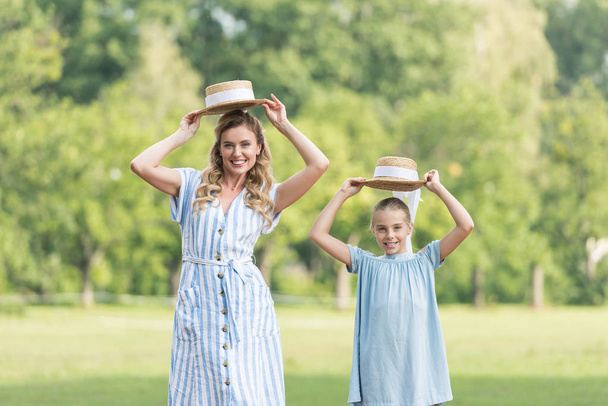 happy mother and adorable daughter posing with straw hats on green lawn - Fotó, kép