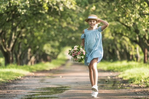 bardzo kobiece dziecko w sukience i słomkowy kapelusz gospodarstwa bukiet kwiatów i chodzenie w ogrodzie - Zdjęcie, obraz