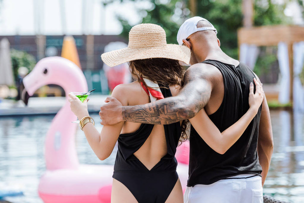 back view of boyfriend and girlfriend hugging and walking near swimming pool with cocktail - 写真・画像