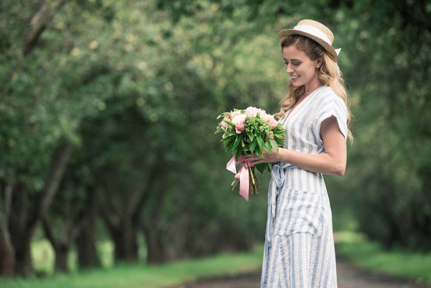 aantrekkelijke lachende vrouw in stro hoed kijken boeket bloemen  - Foto, afbeelding