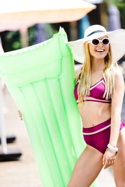 smiling attractive girl in swimsuit and hat standing with inflatable mattress near swimming pool - Photo, image