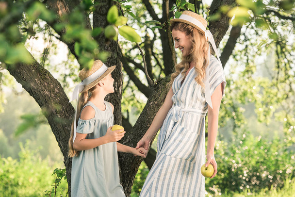 beautiful mother and happy daughter in straw hats with appples looking at each other - Photo, image