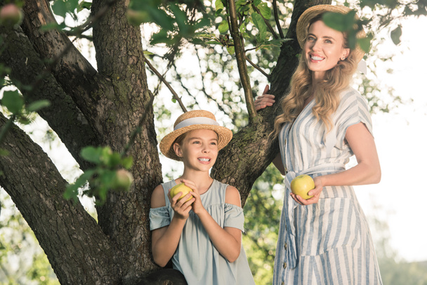 happy mother and daughter in straw hats holding appples and standing near tree - Fotografie, Obrázek