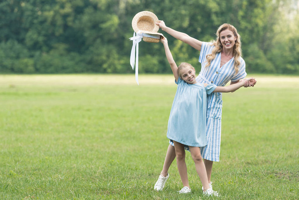 happy mother and daughter posing with straw hats on green lawn - Foto, Imagen