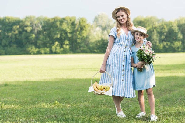 attractive mother and daughter with wicker basket and bouquet posing on green lawn - Photo, Image