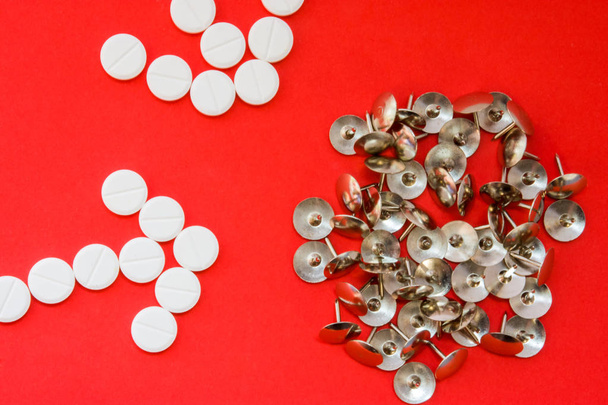 Arrows of pills as a pharmacological agent painkiller and sharp metal thumbtacks as a symbol of pain on red background. Concept photo of action of painkillers or analgesic (anesthetics) in human body - Fotografie, Obrázek