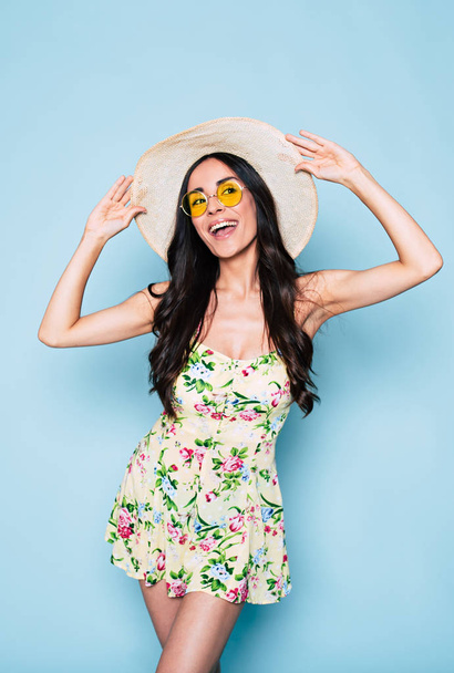 young brunette woman in hat and yellow glasses posing on blue background - Foto, afbeelding