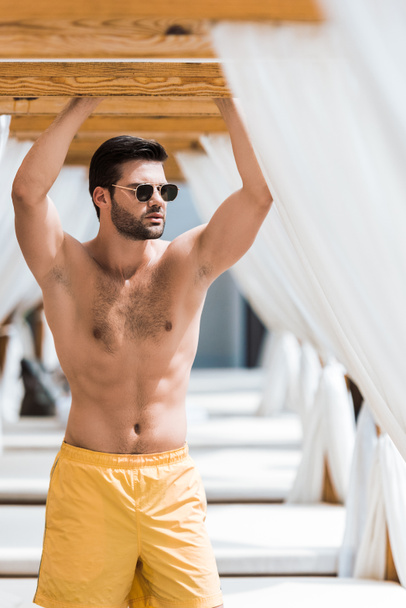 handsome shirtless man in sunglasses standing near sun loungers - Photo, Image