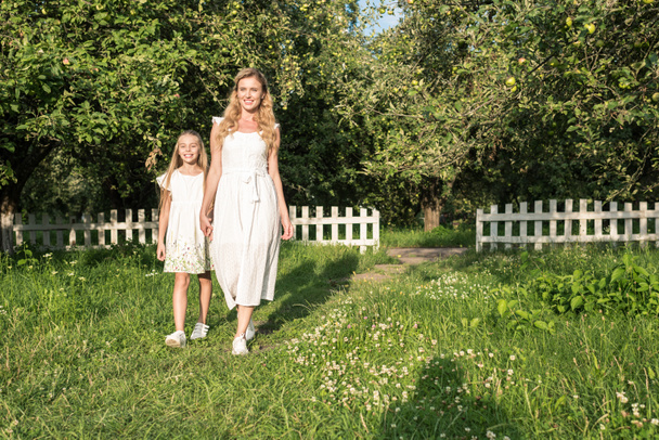 schöne Mutter und Tochter in Kleidern Händchen haltend im Obstgarten mit weißem Zaun - Foto, Bild