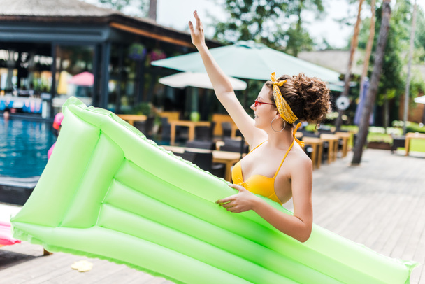 girl in swimsuit walking with inflatable mattress near swimming pool and waving hand - Photo, Image