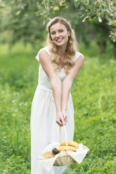bella donna sorridente tenendo cesto di vimini con frutta nel frutteto verde
 - Foto, immagini