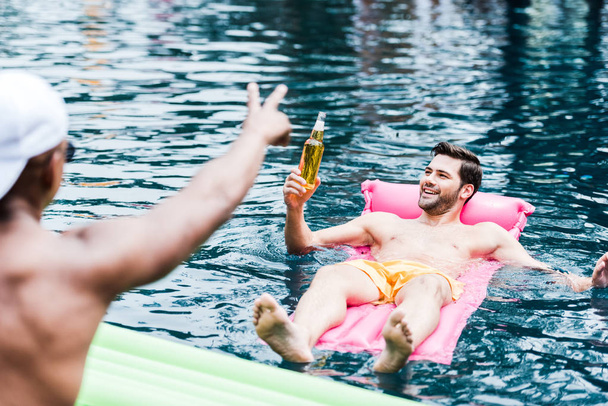笑みを浮かべて男と彼の友人は、プールサイドで指でジェスチャーしながらプールで膨脹可能なマットレスの上に休んでビール - 写真・画像