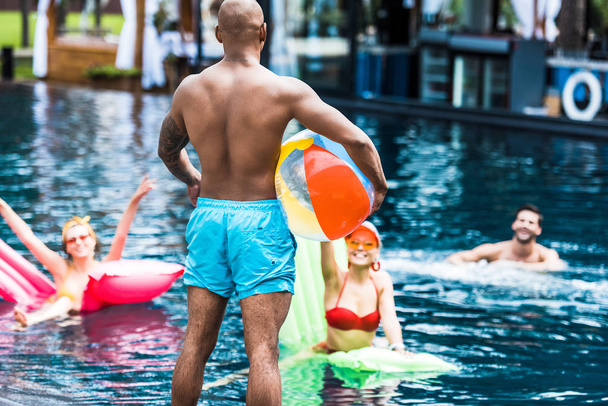 rear view of man standing with swimming ball while his friends in swimming pool with inflatable mattresses - Foto, imagen
