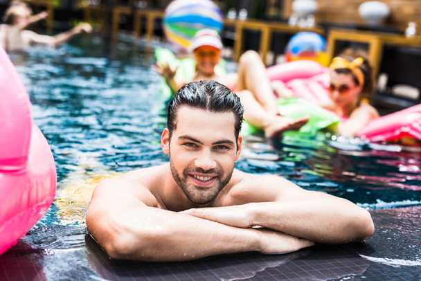 happy young man looking at camera near poolside while his female friends resting on inflatable mattresses in swimming pool - Photo, Image