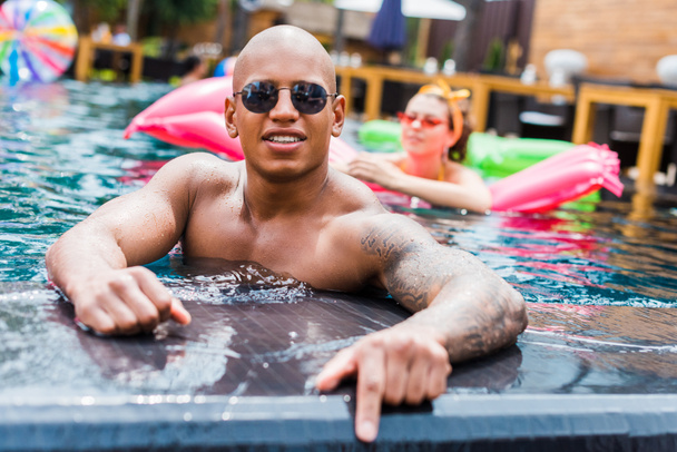 young tattooed man in sunglasses looking at camera while his girlfriend resting on inflatable mattress in swimming pool  - 写真・画像