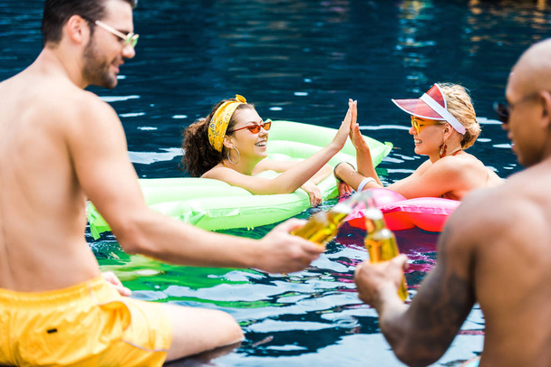 smiling female friends taking high five to each other in swimming pool while male friends clinking by beer bottles  - Photo, Image
