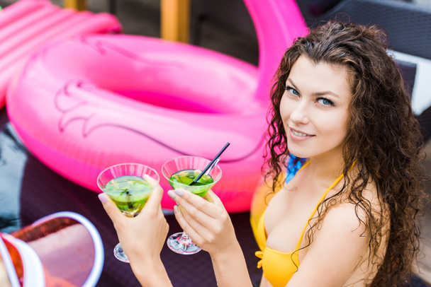smiling woman looking at camera while clinking glasses of cocktails with friend near swimming pool - Photo, Image
