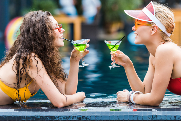 side view of two attractive smiling female friends drinking cocktails on poolside  - 写真・画像