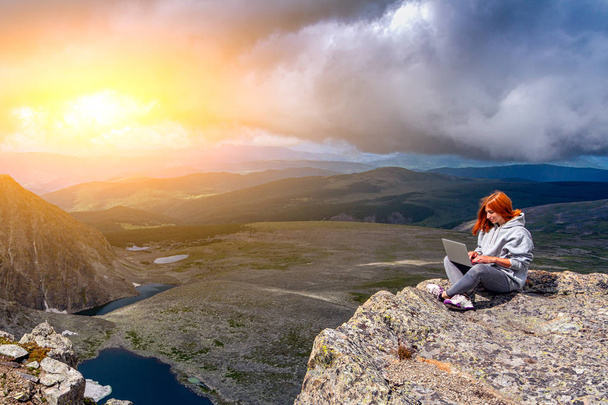Молода туристична жінка працює на ноутбуці, сидячи на вершині кріплення і дивлячись на красивий пейзаж. Фрілансер працює поза офісом. Концепція подорожей та роботи на вулиці
 - Фото, зображення