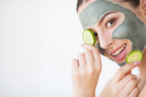 Cuidado de la piel. Mujer joven con máscara de arcilla cosmética sosteniendo pepino en su baño
. - Foto, Imagen