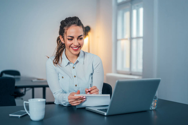Smiling happy businesswoman sitting at desk, looks at laptop screen, making notes. - Foto, Imagem