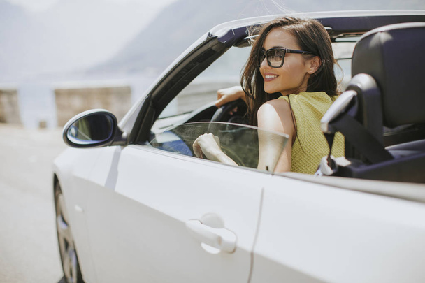 Young woman with sunglasses driving her convertible top automobile on bright sunny day near sea - Photo, Image