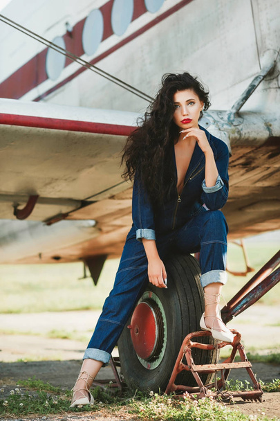 sexy girl with curly hair and in denim overalls near the airplane - Zdjęcie, obraz