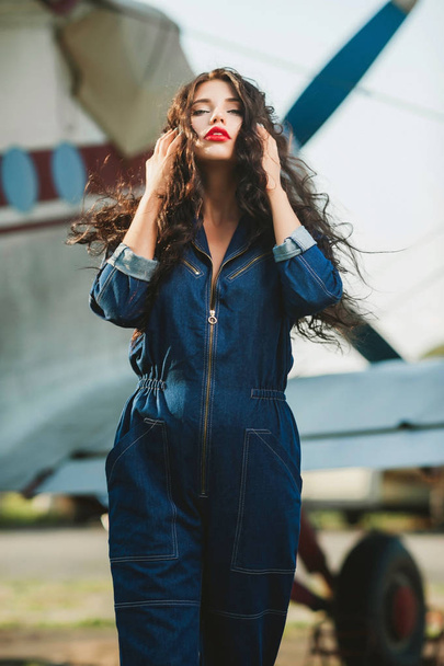 sexy girl with curly hair and in denim overalls near the airplane - Фото, зображення