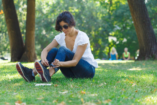 The brunette girl in the park ties up her shoelaces on sneakers. A concept of calm mood and relaxation in a park on the green grass. A middle-aged woman in blue jeans. - Foto, imagen