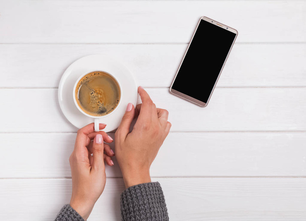 Womans hands holding cup of coffee and smartphone lying nearby - Foto, immagini